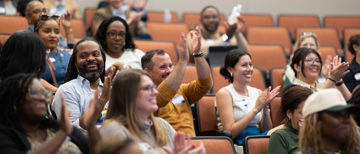 Attendees smile and applaud during a session, several with hands raised.